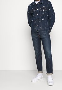 Mörk denimjacka med vitt logotryck över hela ytan, knappstängning och två bröstfickor, kombinerad med något avsmalnande mörkblå jeans och vita sneakers.