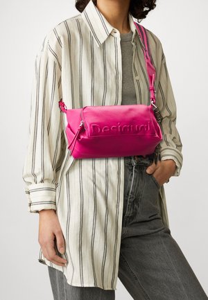 Personne portant une chemise beige à rayures et un jean gris avec un sac bandoulière Desigual rose vif porté en travers de la poitrine.