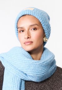 Světle modrý pletený beanie s texturovaným vzorem a krémovou etiketou; doplněné o odpovídající měkký šál, oba vyrobené z silné příze.