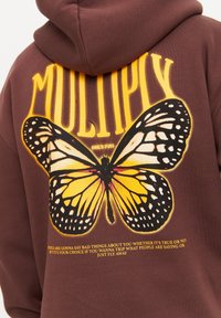 Sudadera marrón con una gran gráfica de una mariposa monarca amarilla y negra. Texto impreso en amarillo arriba y abajo del diseño de la mariposa.