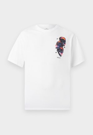 T-shirt blanc avec un graphique vertical de quatre crânes illuminés au néon et le texte "Worldwide Originals" sur la zone gauche de la poitrine.