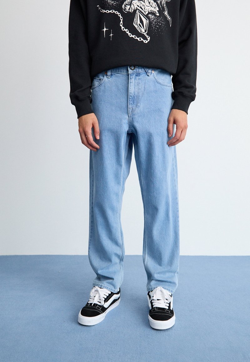 Personne portant un jean droit bleu clair, un sweat-shirt noir avec un motif blanc et des baskets noir et blanc, debout sur un sol bleu.