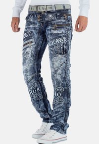 Jeans in denim blu con finitura sbiadita, caratterizzati da loghi ricamati, molteplici tasche con zip e borchie decorative lungo i fianchi.