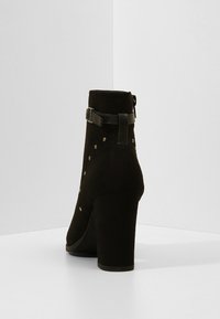 Botín negro de ante hasta el tobillo con tacón cuadrado, que presenta detalles decorativos de tachuelas, un hebilla lateral y una textura suave.
