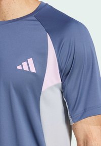 Kraťasový sportovní triko s modrým tělem, růžovými a šedými akcenty, strukturovanou tkaninou a vyraženým logem na hrudi.