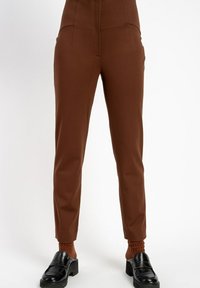 Brązowe, dopasowane spodnie sięgające kostek w zestawieniu z pasującymi teksturowanymi skarpetkami i czarnymi masywnymi loafersami na białym tle.