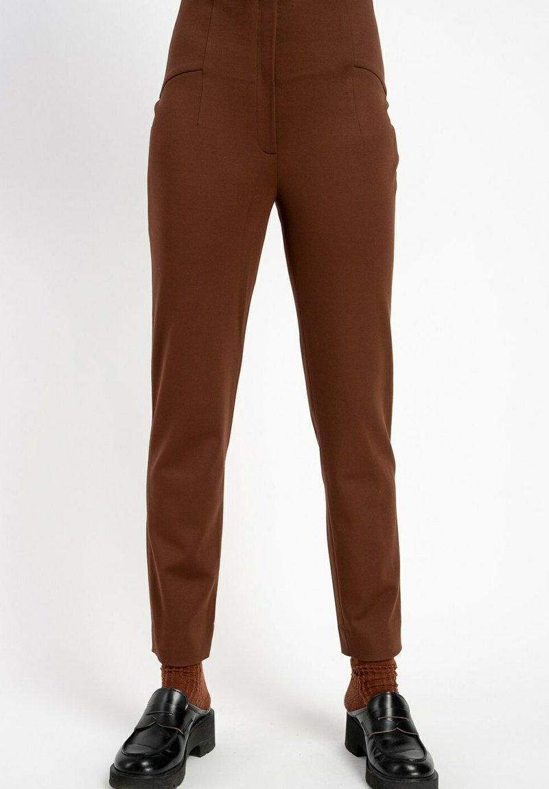 Brązowe, dopasowane spodnie sięgające kostek w zestawieniu z pasującymi teksturowanymi skarpetkami i czarnymi masywnymi loafersami na białym tle.