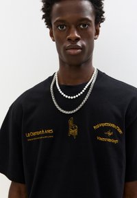 Fekete pamut póló sárga hímzéssel, amely szöveget és egy szék dizájnt ábrázol; gyöngy és ezüstlánc nyakláncokkal rétegezve.