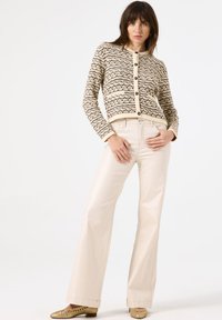 Beiger Strickcardigan mit schwarzem Muster, Rundhalsausschnitt und Knopfverschluss. Kombiniert mit glänzenden cremefarbenen Schlaghosen und goldenen Schuhen.