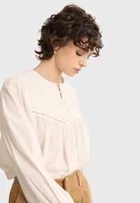 Long-sleeve, ivory blouse with a round neckline, decorative button placket, and gathered bodice. Textured fabric with subtle detailing.
