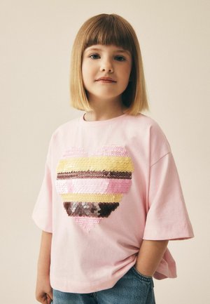 Junges Mädchen mit blondem Bob-Haarschnitt trägt ein pinkes T-Shirt mit einem großen Herz aus mehrfarbigen Pailletten und steht mit einer Hand in der Tasche.