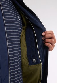 Marineblauwe jas met een rits aan de voorkant, die een gestreept intern shirt en olijfgroene voering onthult. Bevat een logo-opdracht en contrasterende afwerking.