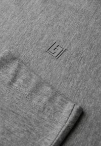 Grauer Stoff mit einer subtilen Textur und einem kleinen, schwarzen quadratischen Logo. Nahtdetails entlang der Kante sichtbar, was das Design des Kleidungsstücks betont.