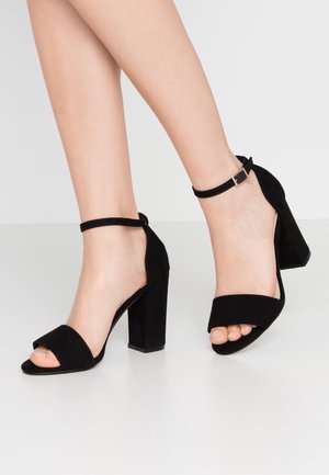 Czarne zamszowe sandały na wysokim obcasie z paskiem wokół kostki, otwartym noskiem i grubym, blokowym obcasem. Gładka faktura z minimalnymi ozdobami.