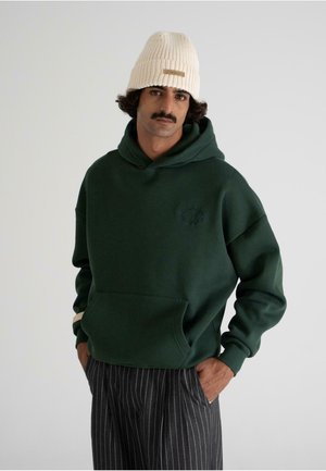 Grüner Hoodie mit einer Fronttasche, der ein dezentes gesticktes Design aufweist. Das Modell trägt eine gerippte cremefarbene Mütze und gestreifte Hosen.