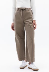 Lichtbruine wijde broek met een elastische tailleband en trekkoorddetail, gemaakt van een gladde stof; gedragen met witte sneakers.