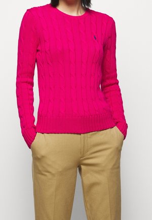 Personne portant un pull torsadé rose vif et un pantalon beige, les mains dans les poches, sur un fond blanc uni.