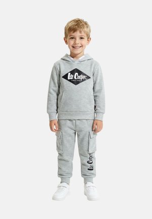 Jeune garçon souriant, portant un sweat à capuche gris Lee Cooper et un pantalon cargo assorti avec des baskets blanches, debout devant un fond blanc uni.