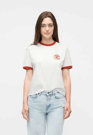 Νεαρή γυναίκα με ίσια καστανά μαλλιά, που φοράει λευκό μπλουζάκι με κόκκινες λεπτομέρειες και λογότυπο στο αριστερό στήθος, συνδυασμένο με ανοιχτόχρωμο τζιν, στέκεται μπροστά σε λευκό φόντο.