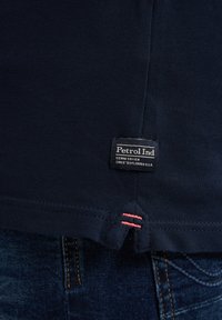 Donkerblauwe gestructureerde stof overhemdzoom met een klein zwart label met de tekst Petrol Ind, bovenop donkerblauwe spijkerbroek met zichtbare stiksels.