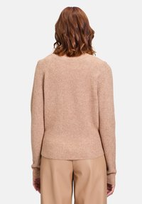 Beige geripptes Strickoberteil mit rundem Ausschnitt und langen Ärmeln, ausgestattet mit einer taillierten Silhouette und weichem Material. Getragen mit beigen Hosen.
