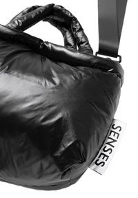 Schwarze, gepolsterte Schultertasche mit glänzender Textur, ausgestattet mit zwei Griffen und einem verstellbaren Riemen. Weißes Etikett mit der Aufschrift "SENSES" an der Seite.