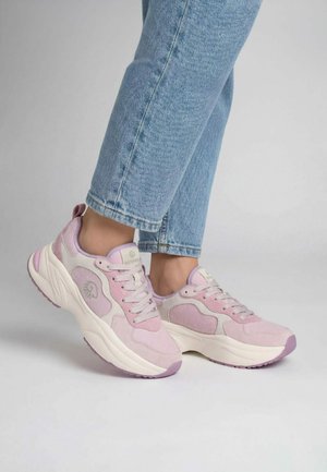 Osoba nosząca jasnoniebieskie dżinsy i pastelowo różowe oraz beżowe masywne sneakersy stojąca na prostej, jasnoszarej podłodze.