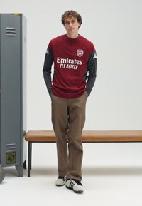 Veste de sport rouge Arsenal avec un col montant et des manches grises, pantalon marron, et chaussures noires et blanches, debout à côté d'un casier et d'un banc.