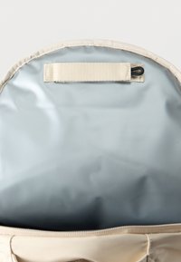 Ανοιχτό μπεζ σακίδιο πλάτης που δείχνει το άδειο εσωτερικό σε ανοιχτό γκρι χρώμα με μπεζ λουρί κοντά στην άνω άκρη.