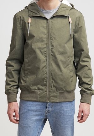 Man draagt een olijfgroene hoodie met rits en witte trekkoorden, gecombineerd met lichtblauwe jeans, staand voor een neutrale achtergrond.