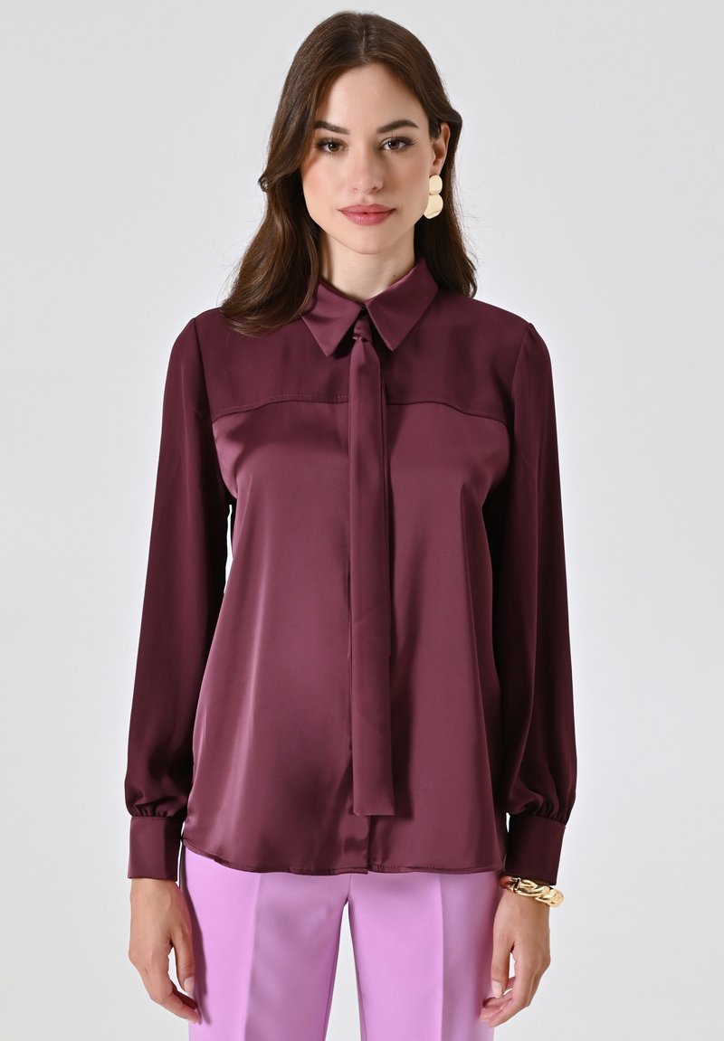 Blusa in satin bordeaux con colletto, dettaglio dei lacci e maniche lunghe. Texture liscia e leggero lucido, abbinata a pantaloni rosa chiaro.