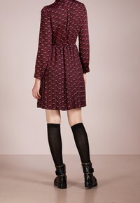 Abito borgogna con motivo circolare, vita cinchata e maniche lunghe. Abbinato a calze nere al ginocchio e stivaletti alla caviglia.