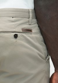 Hellgraue Hose mit glatter Textur, die eine Logo-Patch am Bund und einen Knopfverschluss an der vorderen Tasche aufweist.