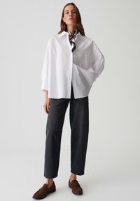 Femme portant une chemise blanche oversize à boutons, un foulard noir à motifs autour du cou, un jean noir à jambes droites, et des mocassins marron, debout avec une main dans la poche.