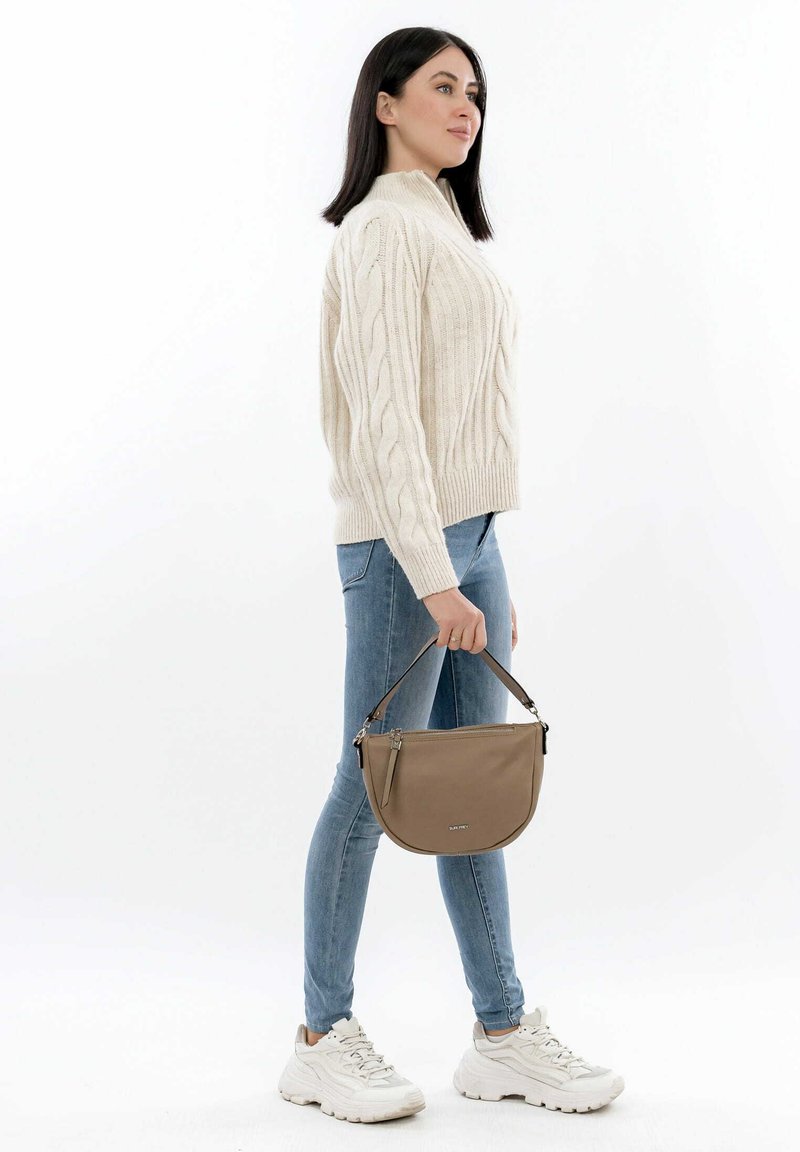 Maglione lavorato a maglia beige, jeans skinny blu e sneakers bianchi. Tiene in mano una borsa curva marrone con una tracolla corta e dettagli con zip.