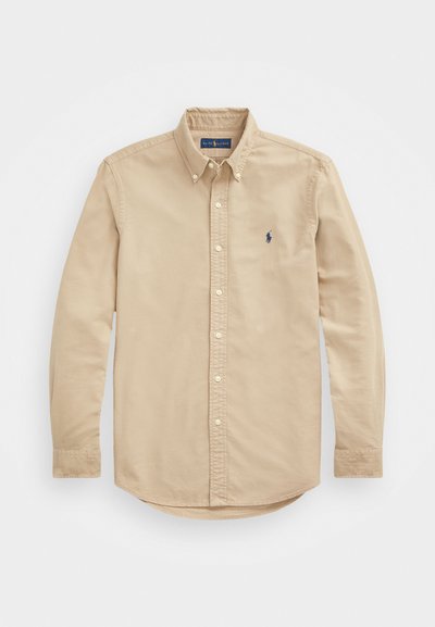 Ljusbeige knäppt skjorta med spetsig krage, långa ärmar och en blå broderad logotyp på vänster bröst. Mjuk bomullstyg.