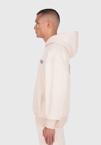 Mężczyzna z krótkimi włosami w kremowej bluzie z kapturem i jasnych spodniach, stojący w profilu na jednolitym tle.