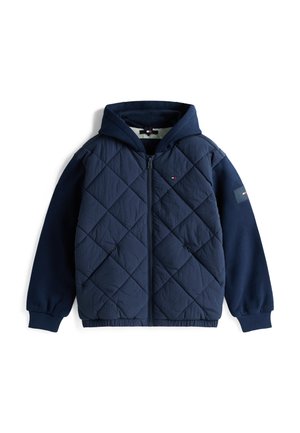 Veste matelassée bleu marine avec capuche, présentant des motifs en losanges, des poignets côtelés et une patch logo sur la manche. Fermeture éclair à l'avant et poches incluses.