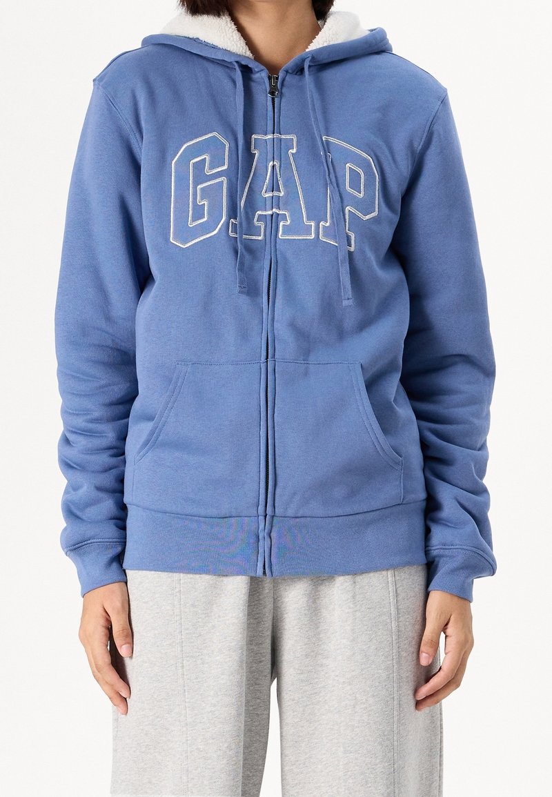 Personne portant un sweat à capuche bleu zippé avec doublure en polaire blanche et grandes lettres blanches « GAP » à l'avant, associé à un pantalon de survêtement gris clair.