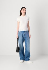 T-shirt bianca aderente, jeans a gamba larga blu, sneaker bianche e borsa a tracolla nera. Design semplice con un piccolo logo sulla maglietta.