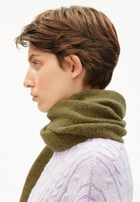 Persoon met kort bruin haar, gekleed in een groene wollen sjaal en een lichtpaarse kabelgebreide trui, die naar links kijkt op een witte achtergrond.