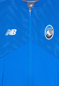 Blå pustende jakke med glidelås, New Balance-logo på venstre bryst og Atalanta 1907 fotballklubb-emblem på høyre bryst.