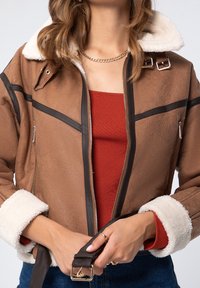 Veste en faux daim marron avec un col en fausse fourrure blanc, des finitions noires, une fermeture éclair à l'avant et une ceinture décorative. Portée par-dessus un top rouge à côtes.