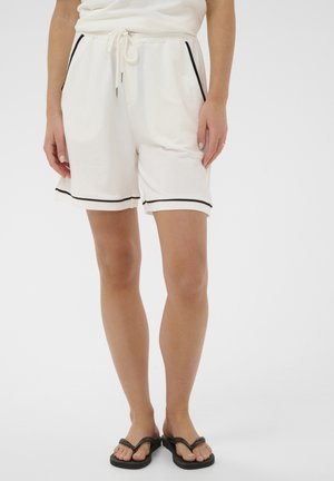 Kobieta w białych szortach ze sznurkiem i czarnym obszyciem oraz czarnych japonkach, stojąca na tle jasnego, jednolitego tła.