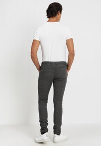 Muž stojící s rukama v kapsách, oblečený v bílém tričku, tmavě šedých slim-fit kalhotách a bílých teniskách, na jednobarevném světlém pozadí.