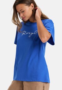 Camiseta de algodón azul con mangas cortas, con logo en cursiva blanco "Roxy" y acento floral. Corte estándar con una textura suave.
