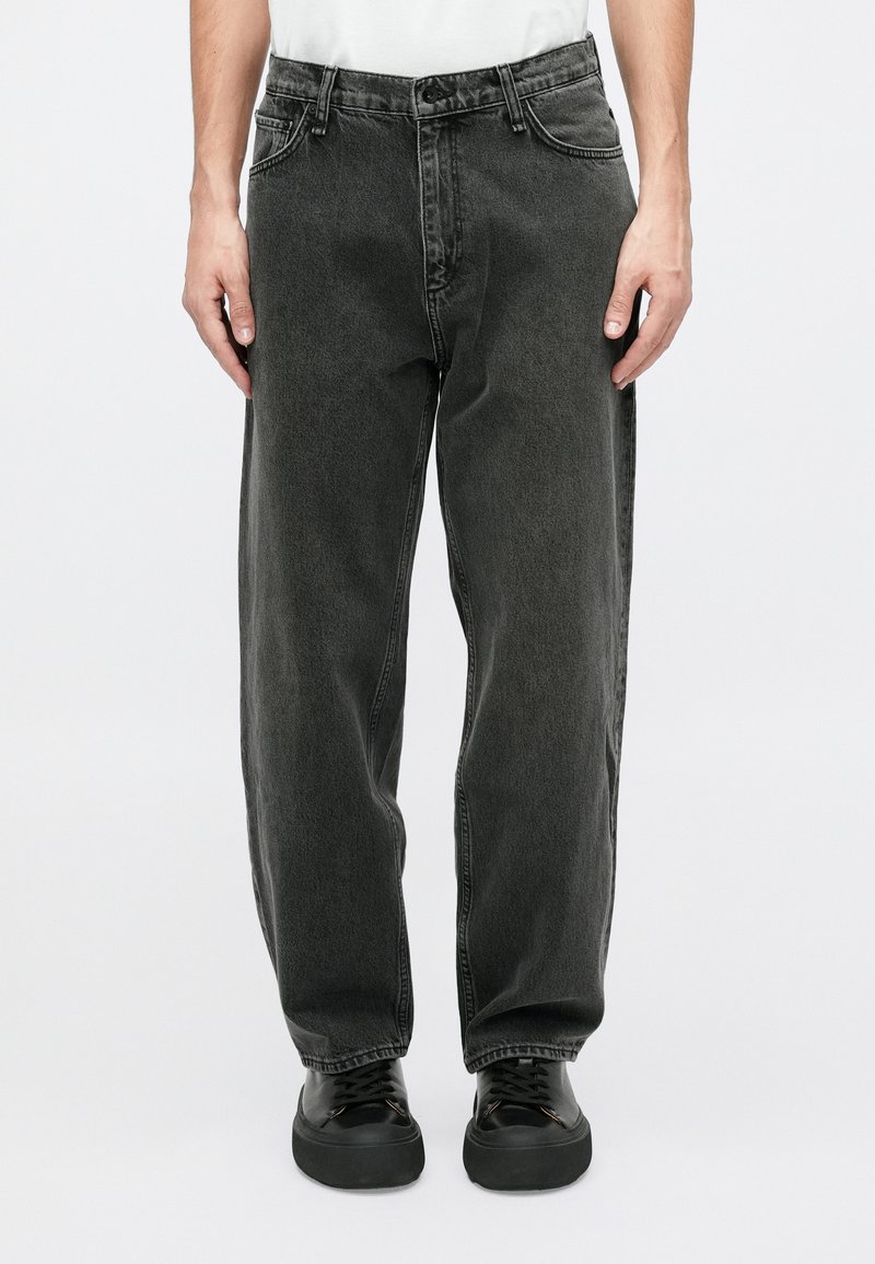 Person iført løstsiddende, sort, vaskede jeans og sorte sneakers, stående imod en ensfarvet baggrund.