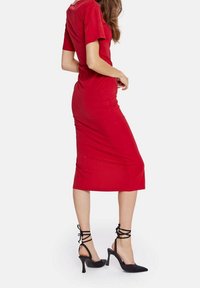 Rotes Midi-Kleid mit kurzen Ärmeln, figurbetonter Silhouette und seitlichem Schlitz. Kombiniert mit schwarzen, geflochtenen High-Heels mit spitzer Zehenpartie.