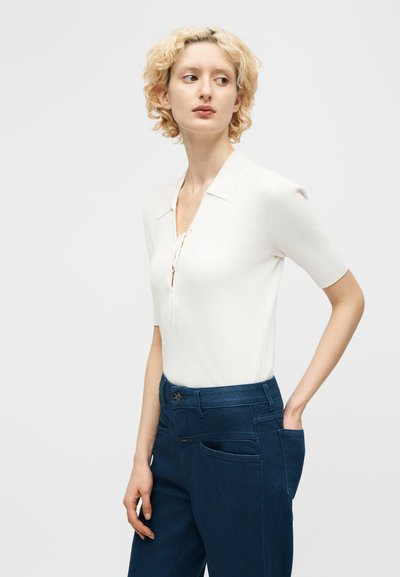 Ξανθιά γυναίκα με λευκή πλεκτή μπλούζα κοντών μανικιών με κορδόνια στο μπροστινό μέρος, φοράει σκούρο μπλε ψηλόμεσο παντελόνι, κοιτάζοντας αριστερά της.