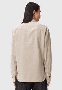 Maglietta beige a maniche lunghe in cotone, con polsini abbottonati, taglio dritto e colletto classico, mostrata di spalle.